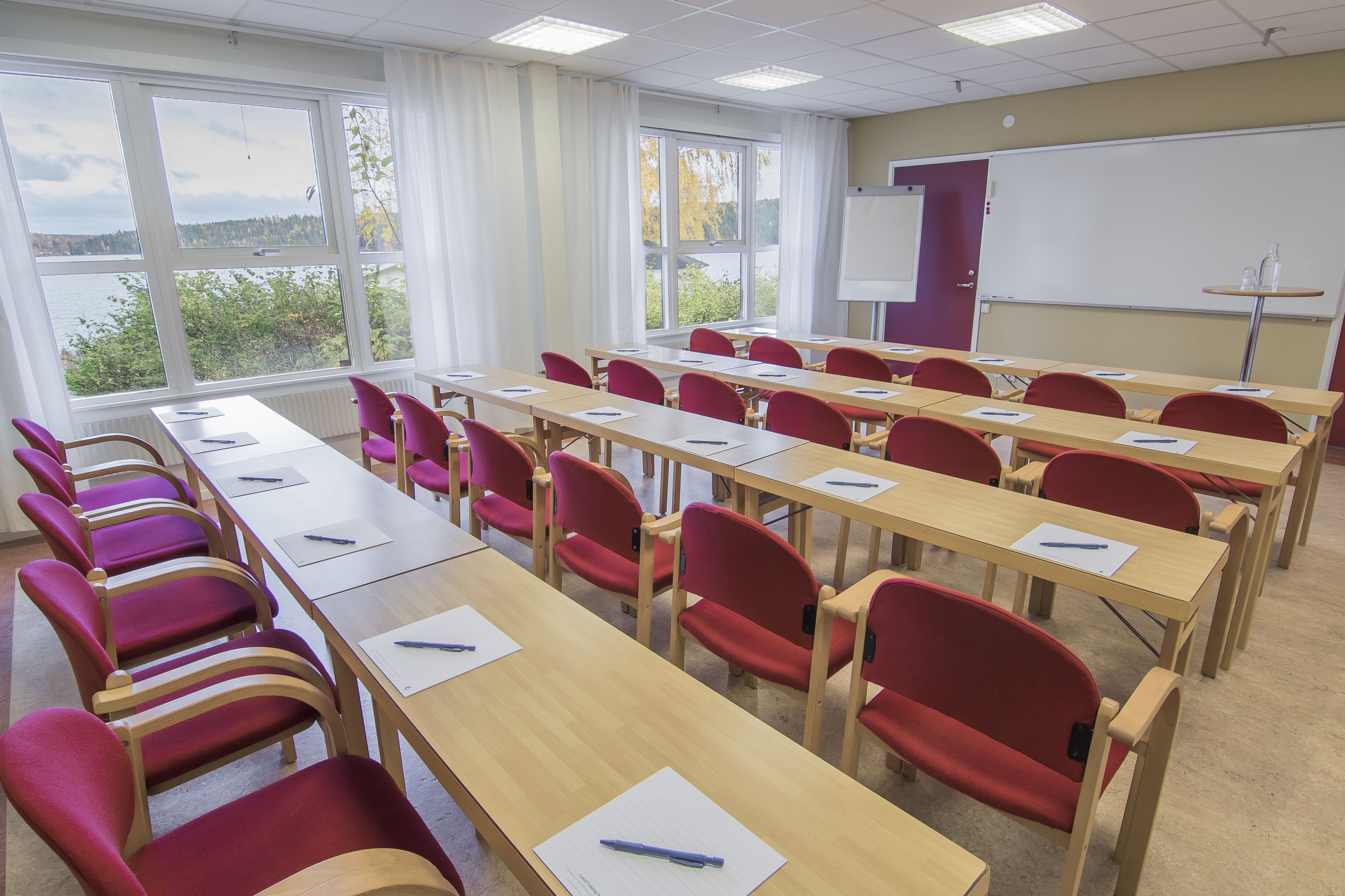 A classroom with rows of desks and chairs, large windows, and a whiteboard.
