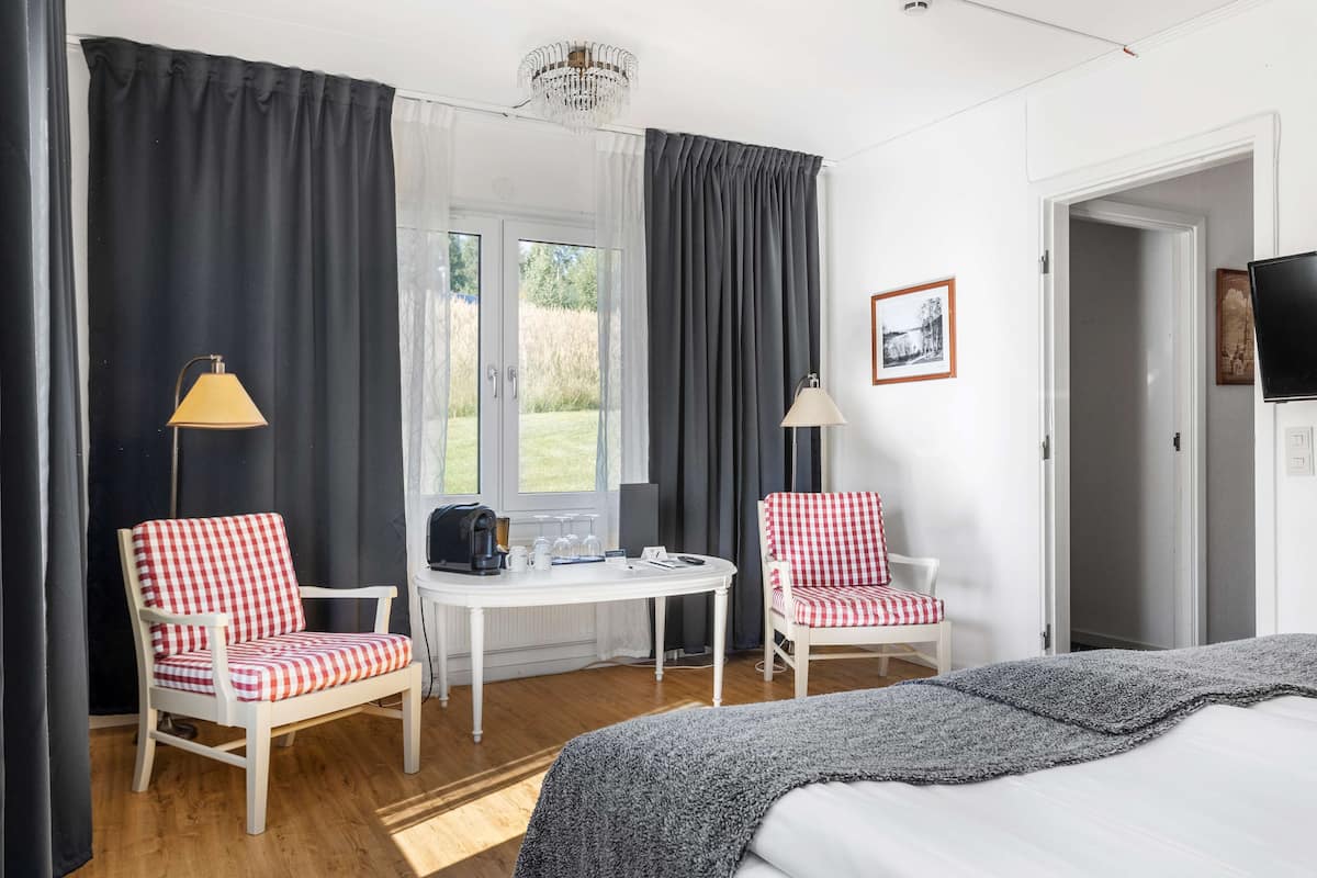A room with a bed, two chairs, a small table with a kettle and cups, a television, and a view of the outdoors.