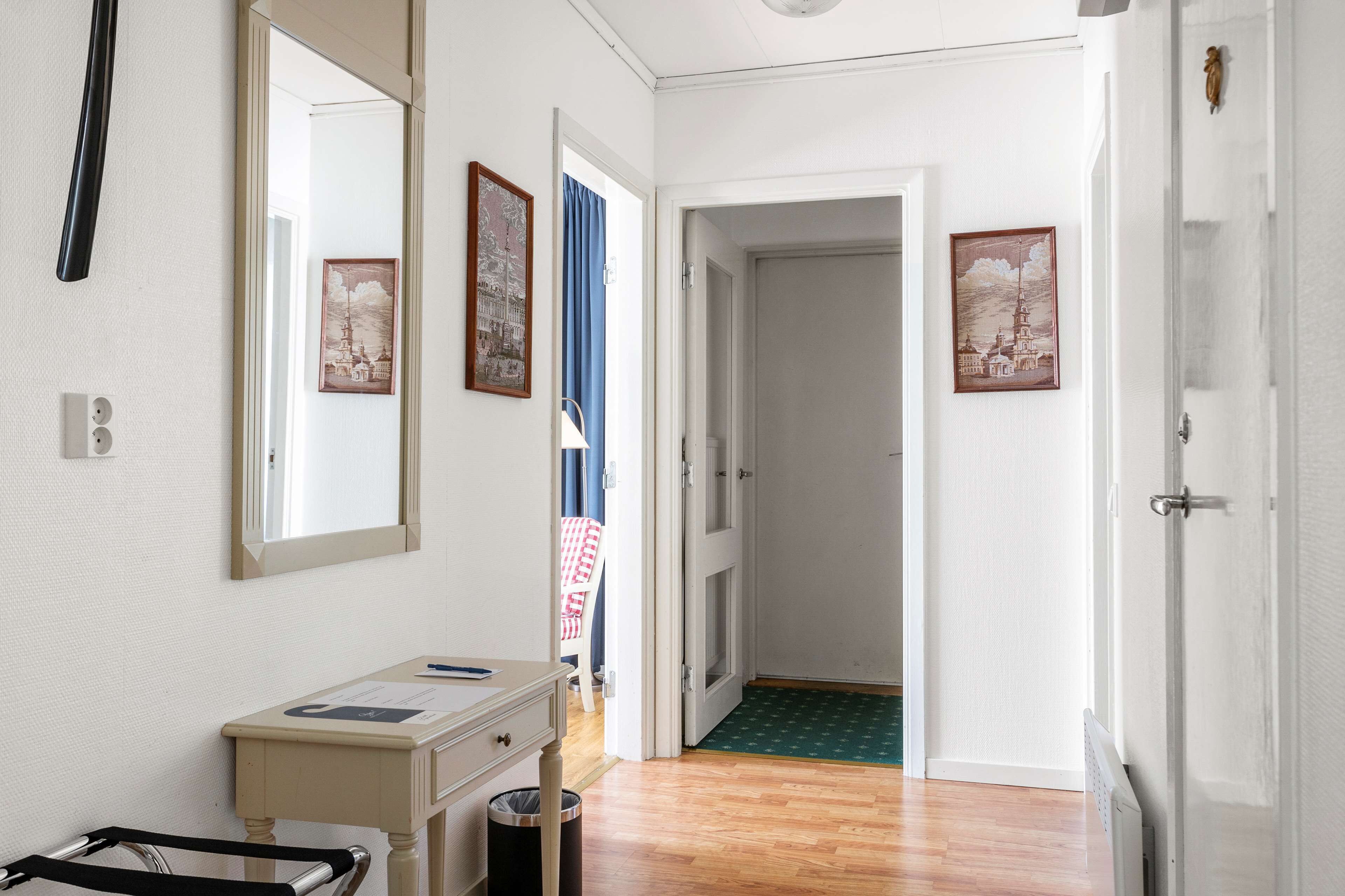 A hotel room with a wooden floor, a bedside table with a lamp, a mirror, and two framed pictures on the wall.