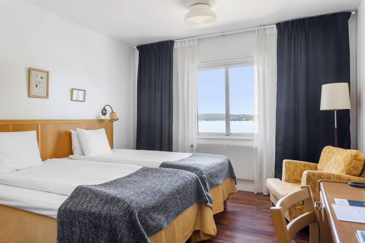 A hotel room with two beds, a wooden headboard, a dining table with a chair, a lamp, and a view of the sea through large windows.