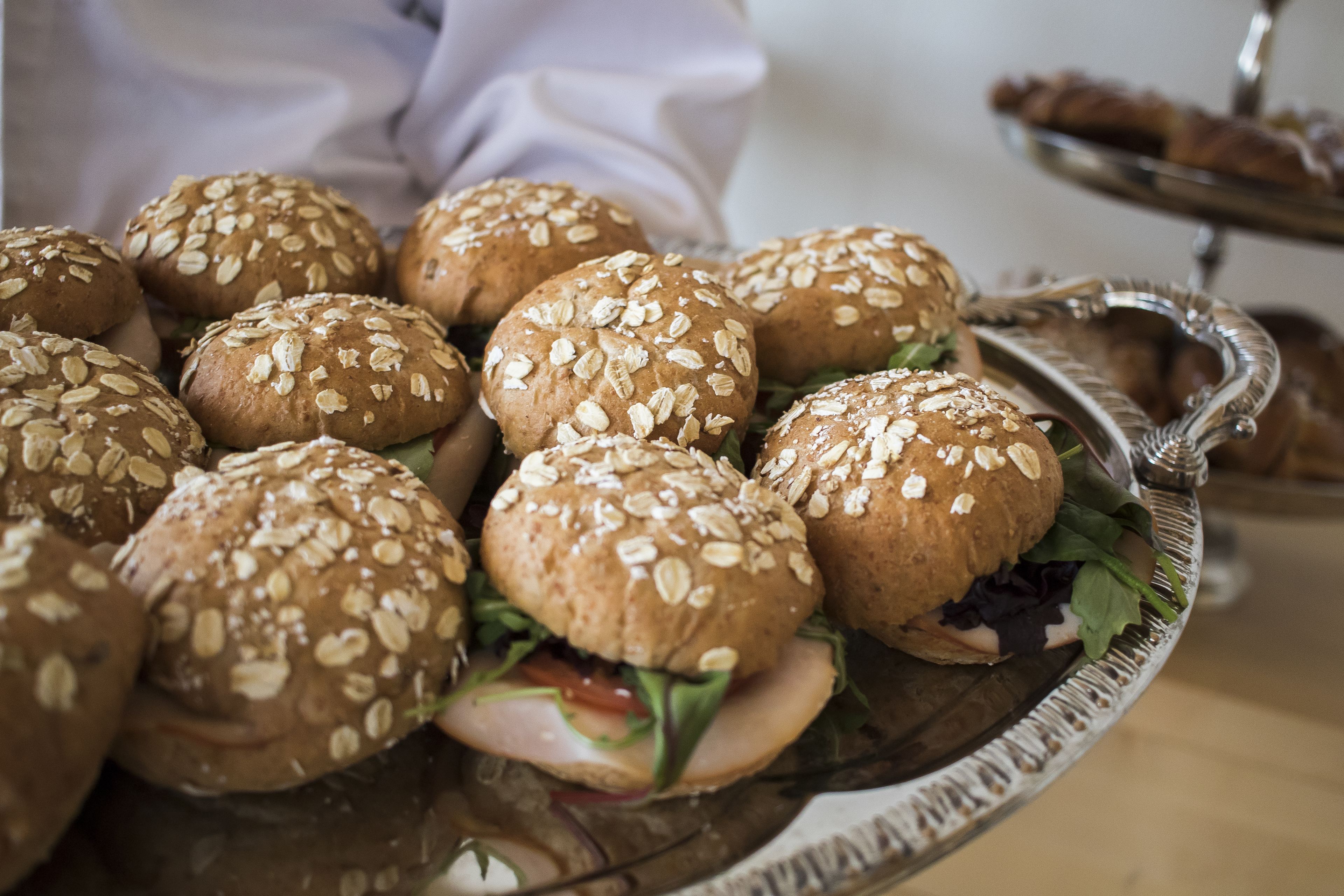 A tray of sandwiches with sesame seeds on buns, a salad, and a dessert tray in the background.