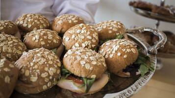 A tray of sandwiches with sesame seeds on buns, a salad, and a dessert tray in the background.