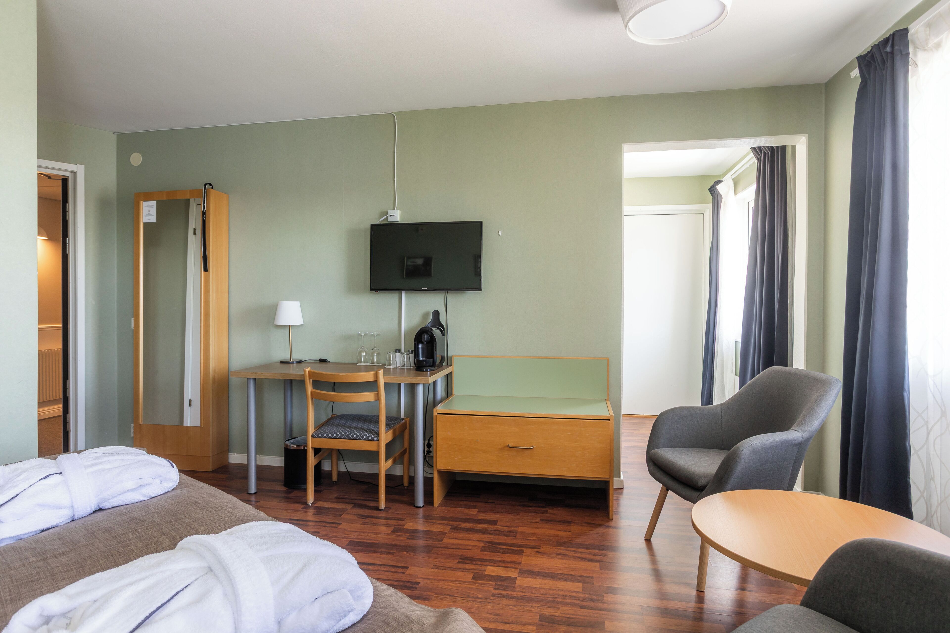 A hotel room with a bed, a desk with a chair, a television, a coffee maker, and a window with curtains.