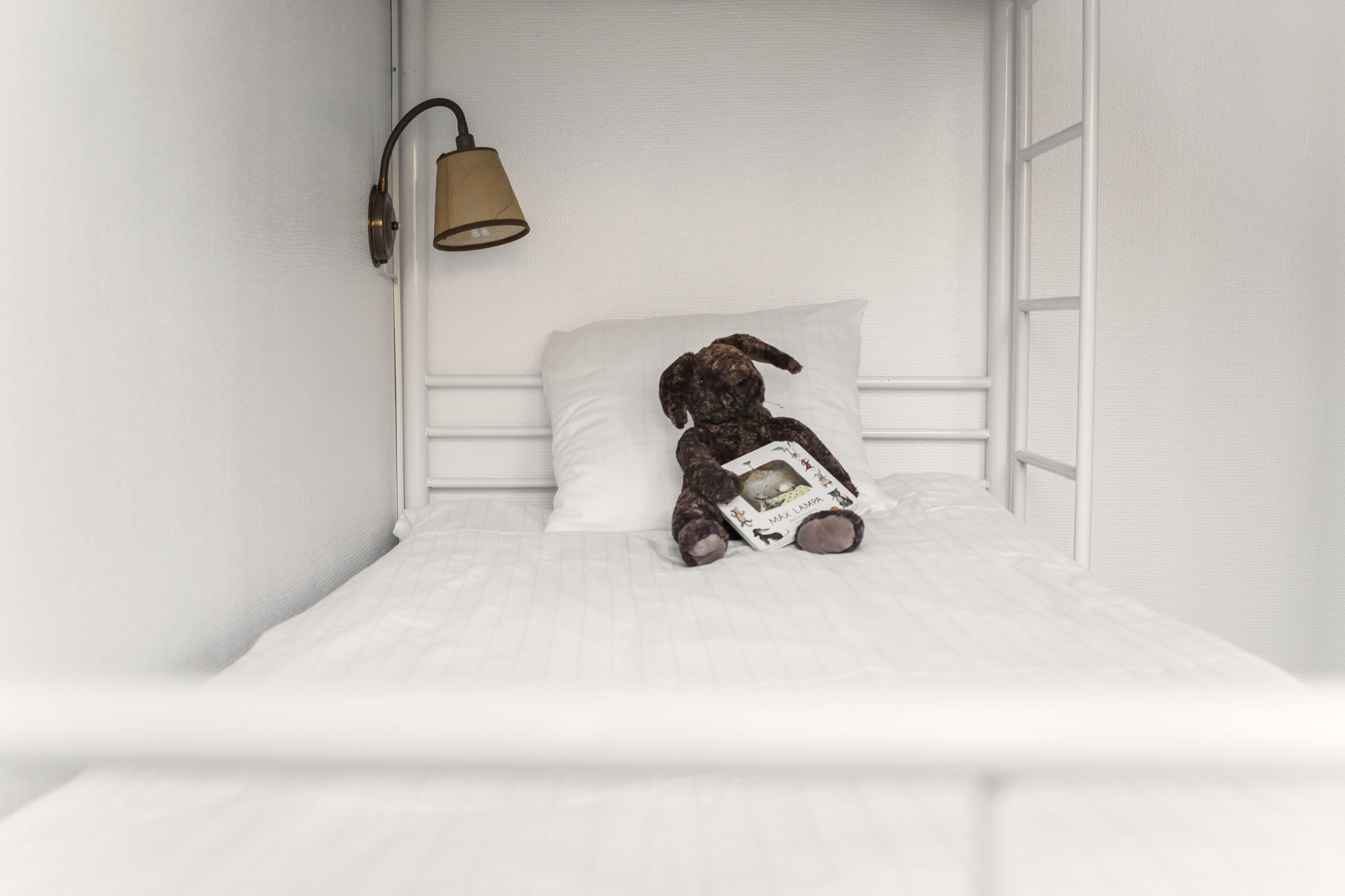 Una cama con colchón y cabecera blancos, una almohada blanca y un osito de peluche sosteniendo un libro.