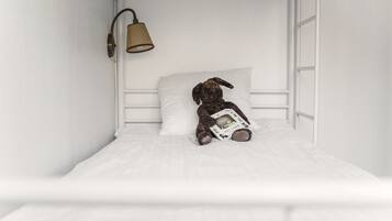Una cama con colchón y cabecera blancos, una almohada blanca y un osito de peluche sosteniendo un libro.