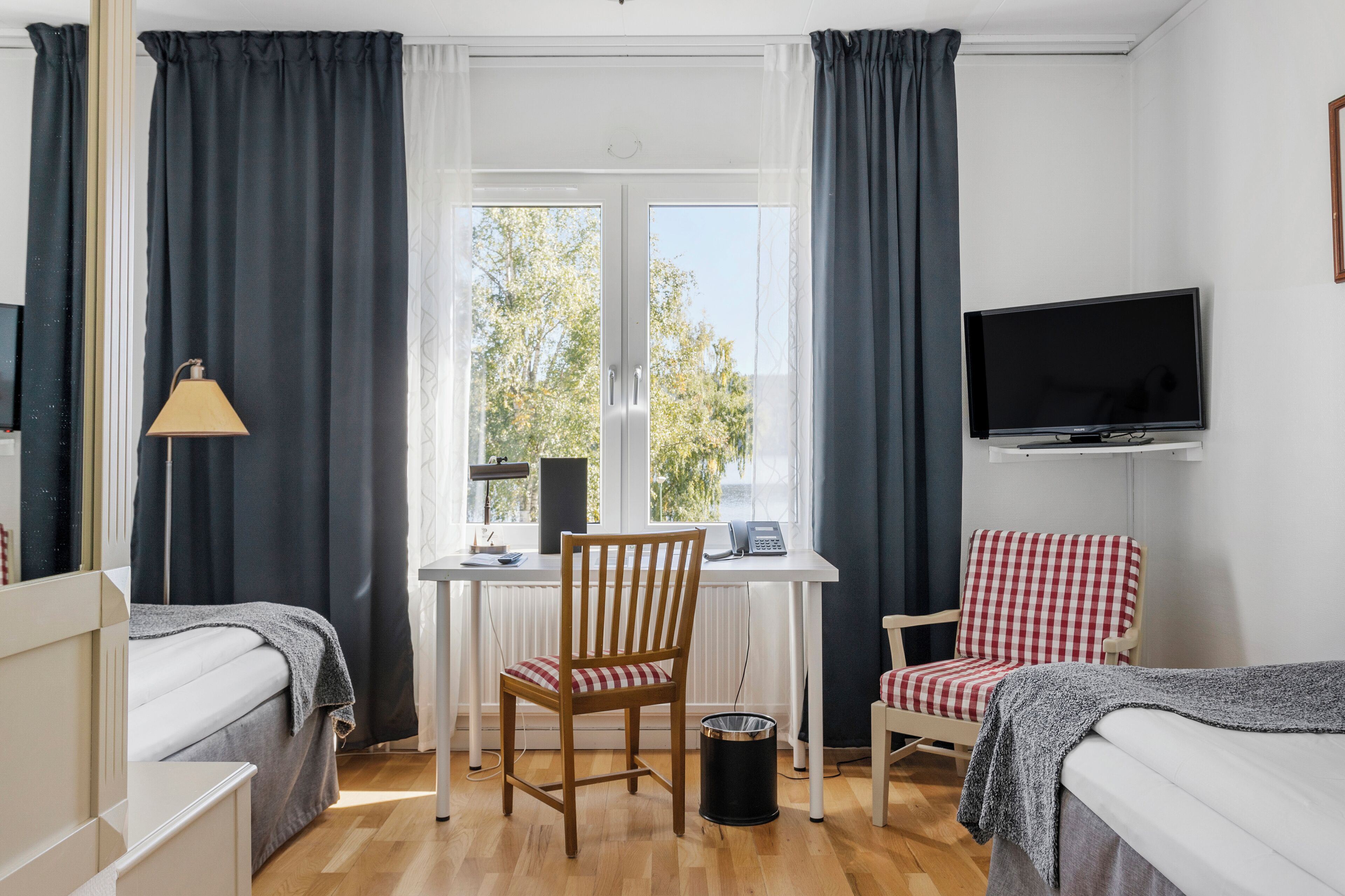 Ein Zimmer mit zwei Betten, einem Schreibtisch mit Stuhl, einem Fernseher und einem Fenster mit Vorhängen.