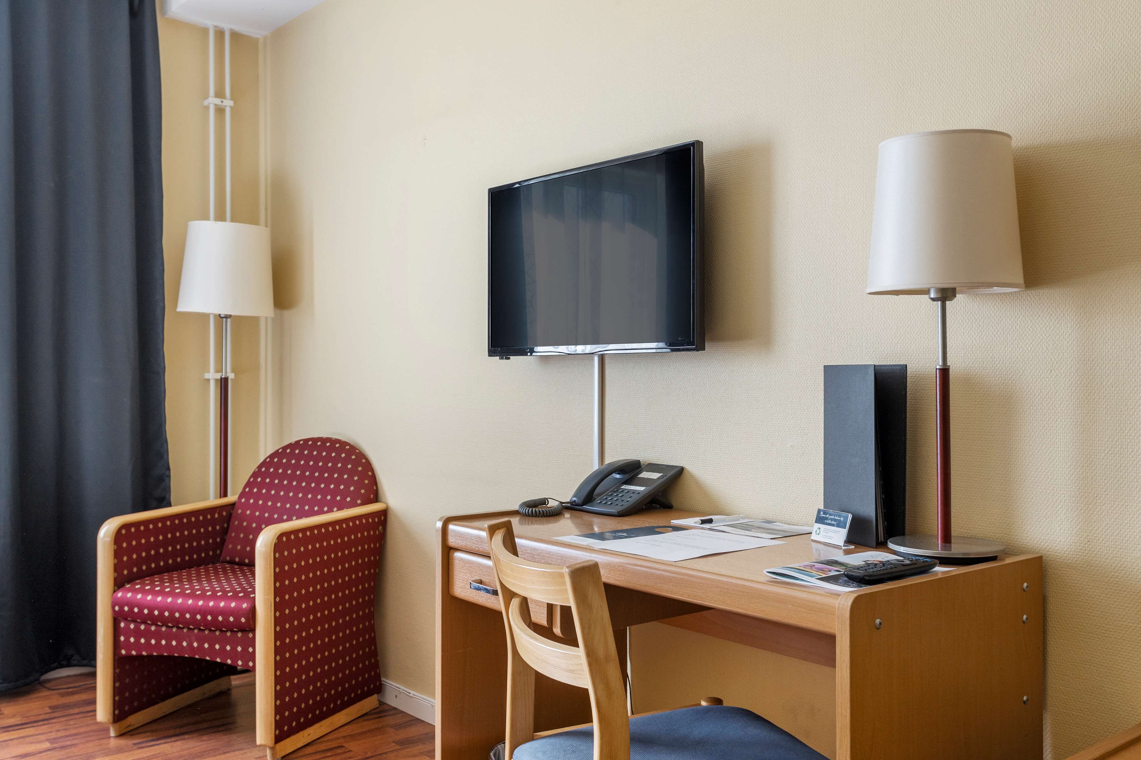 Une chambre d’hôtel avec un bureau en bois, une chaise à motifs rouges, un téléviseur fixé au mur et deux lampadaires.