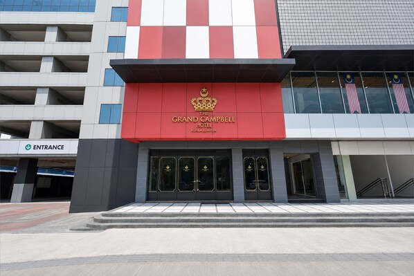 L’entrée d’un hôtel avec une façade rouge et blanche, un emblème représentant une couronne, et l’inscription « The Grand Campbell Hotel Kuala Lumpur ».