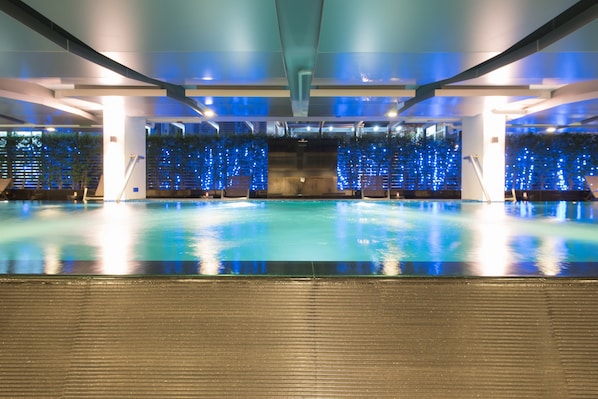 A large indoor swimming pool with a modern design, surrounded by seating areas and a decorative wall with blue lights.
