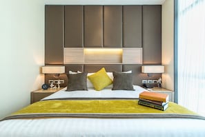 A hotel room with a large bed, a headboard with built-in lighting, bedside tables with lamps, and a window with curtains.