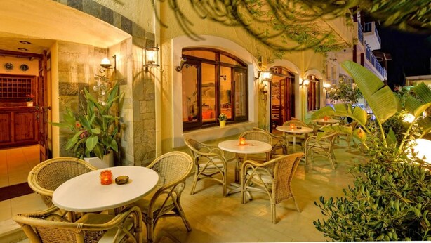 An outdoor patio area with wicker furniture and tables, surrounded by plants and a building with a stone facade. - Floral Hotel (Hersonissos)