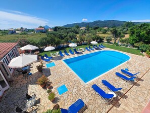A swimming pool with lounge chairs and umbrellas, surrounded by stone tiles and potted plants. - Elena Pool Apartments (Corfu)