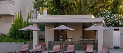 Un área junto a la piscina con sillones de descanso y sombrillas, un bar y una piscina.