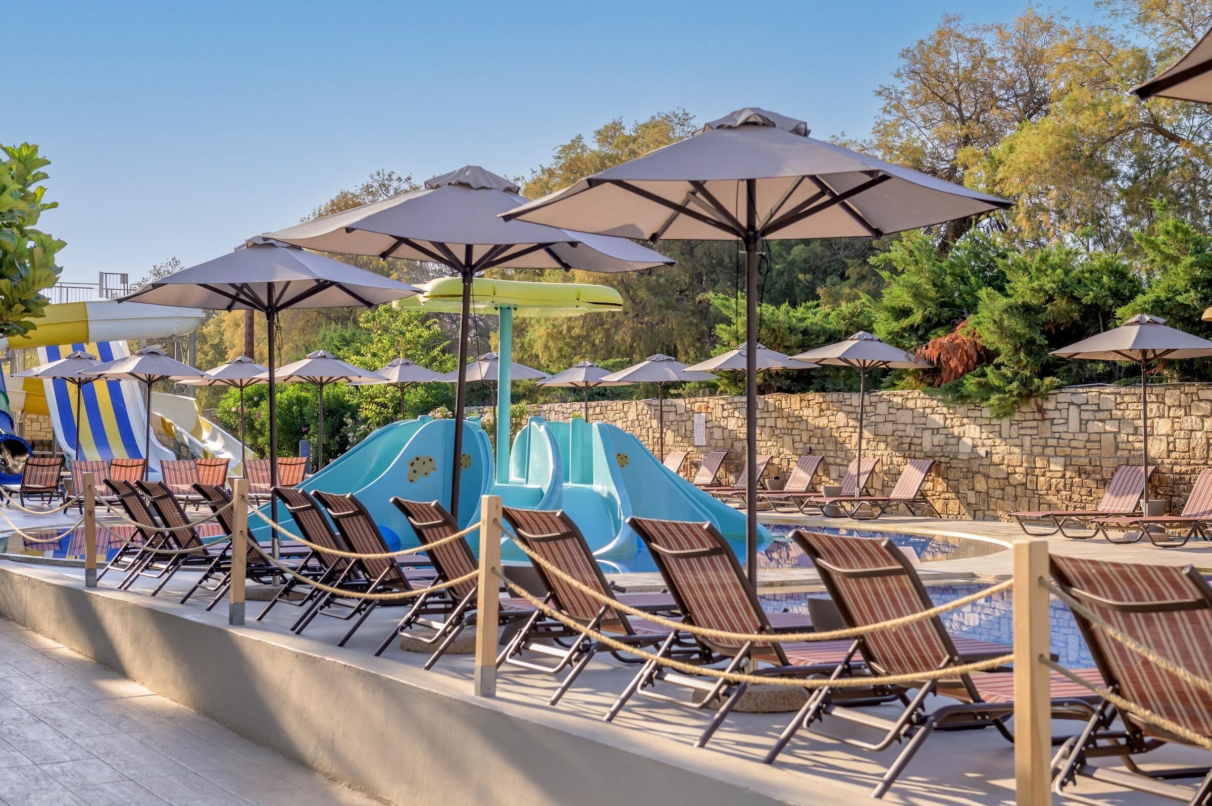 A poolside area with lounge chairs and umbrellas, a water slide, and a stone wall.