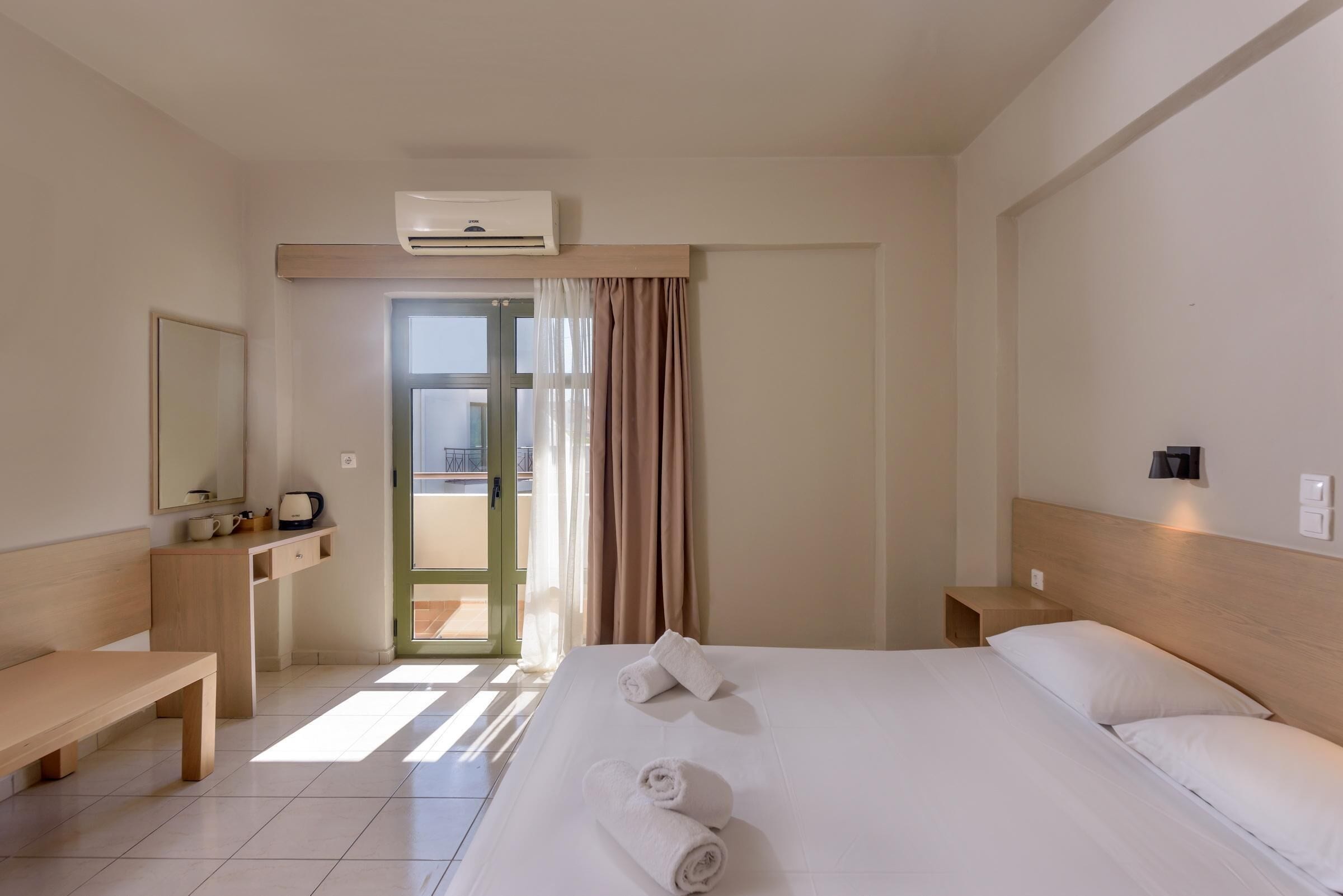 A hotel room with a bed, bedside tables, a desk, a chair, an air conditioning unit, and a view through the door.