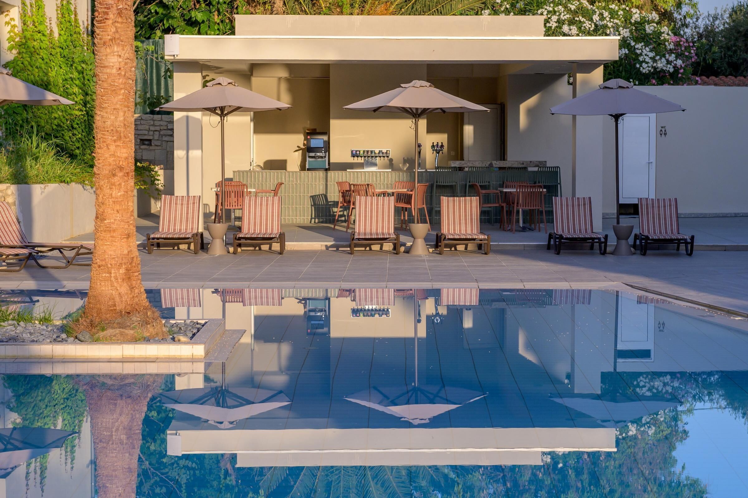Zona junto a la piscina con sillones de descanso y sombrillas, un bar y una piscina reflectante.