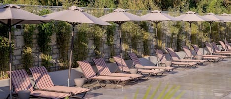 Een zwembadterras met ligbedden en parasols, omgeven door groen en stenen muren.