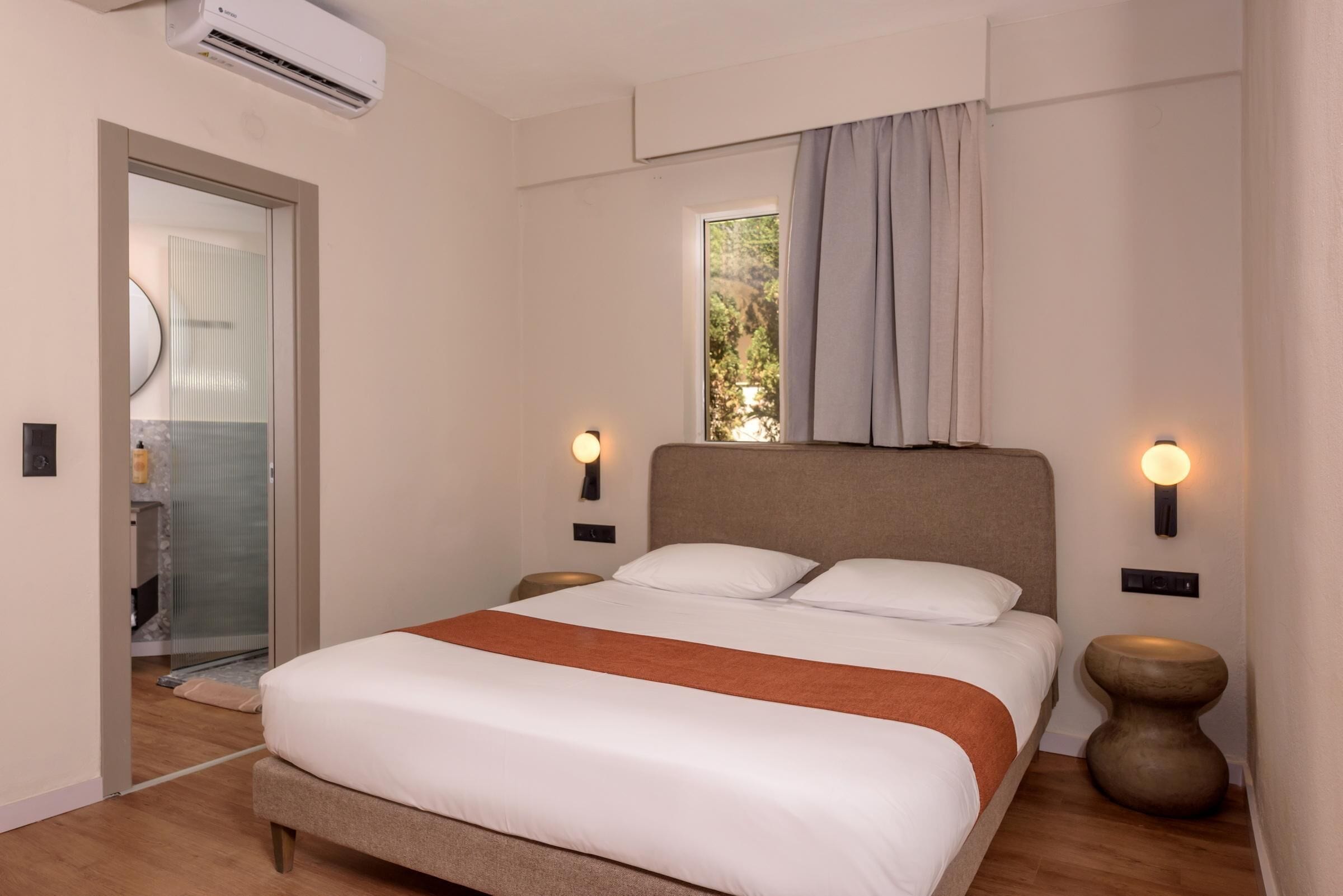 A bedroom with a bed, two bedside tables, an air conditioning unit, a window with curtains, and a door to a bathroom.