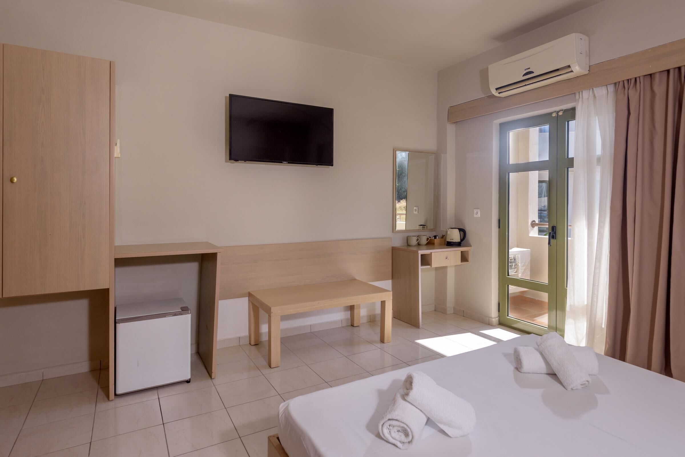 Une chambre d’hôtel équipée d’un lit, d’une télévision, d’un mini-réfrigérateur, d’un bureau, d’une banquette, d’un miroir et d’une porte donnant sur un balcon.