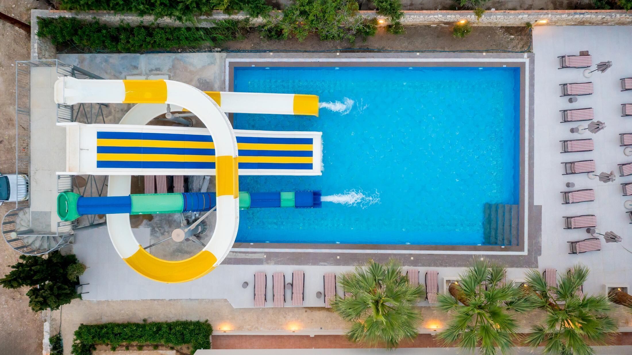 Vue aérienne d’une piscine avec un toboggan, des chaises longues et des palmiers.