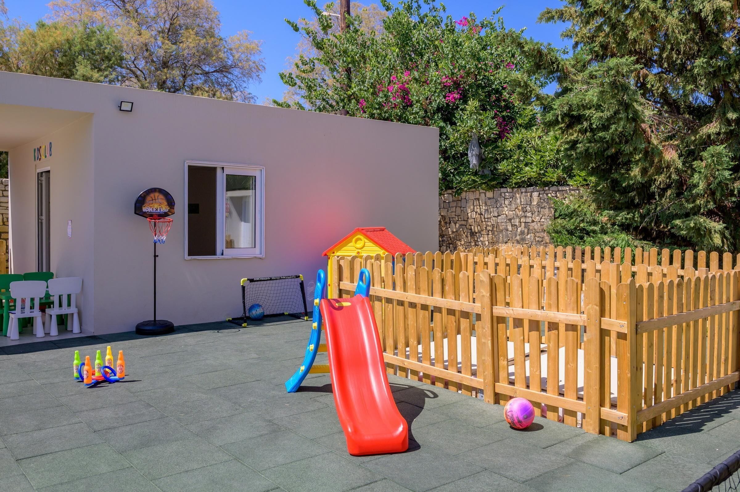 A playground with a red slide and a climbing net, surrounded by a wooden fence and trees.