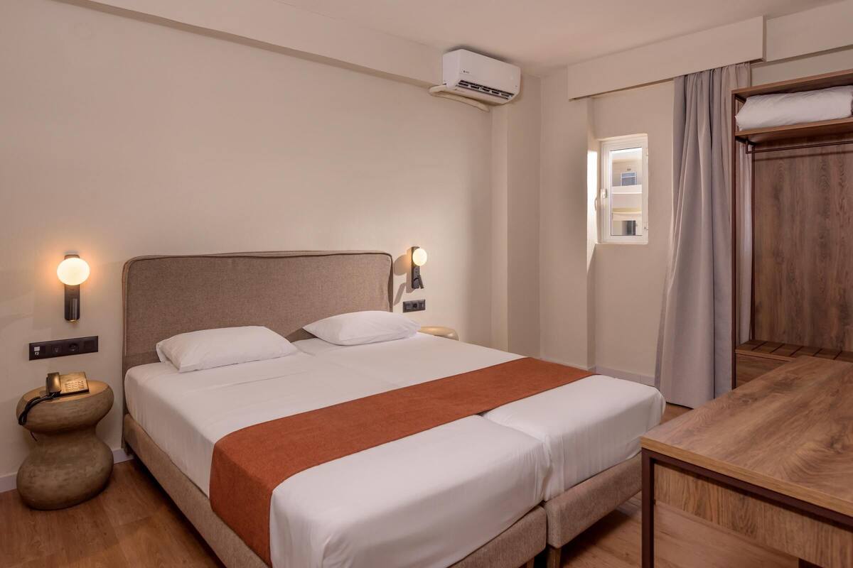 A hotel room with a bed, bedside lamps, an air conditioning unit, a wooden nightstand, a wooden desk, and a window with curtains.