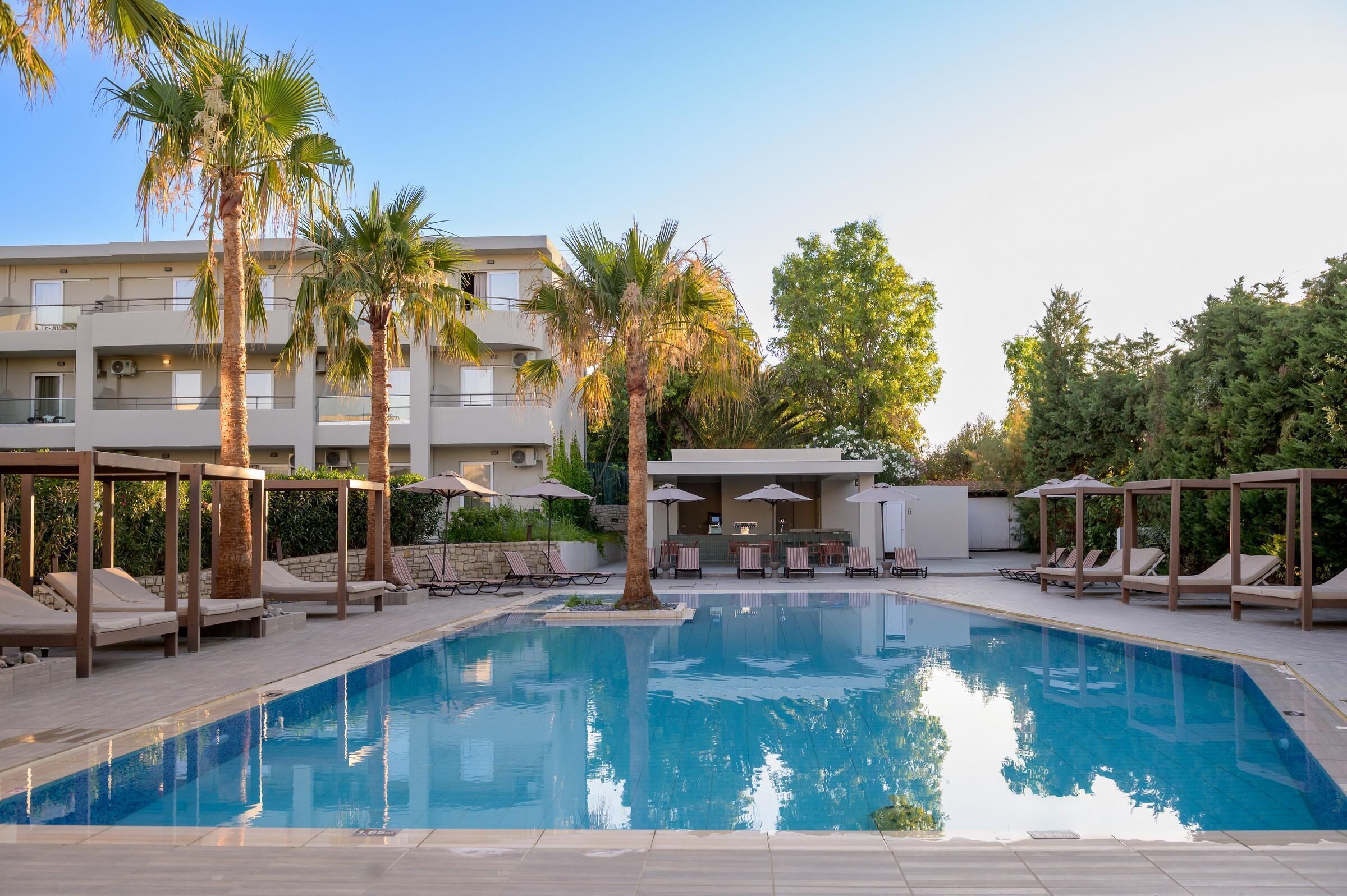 A swimming pool with lounge chairs and umbrellas, surrounded by palm trees and a multi-story building.