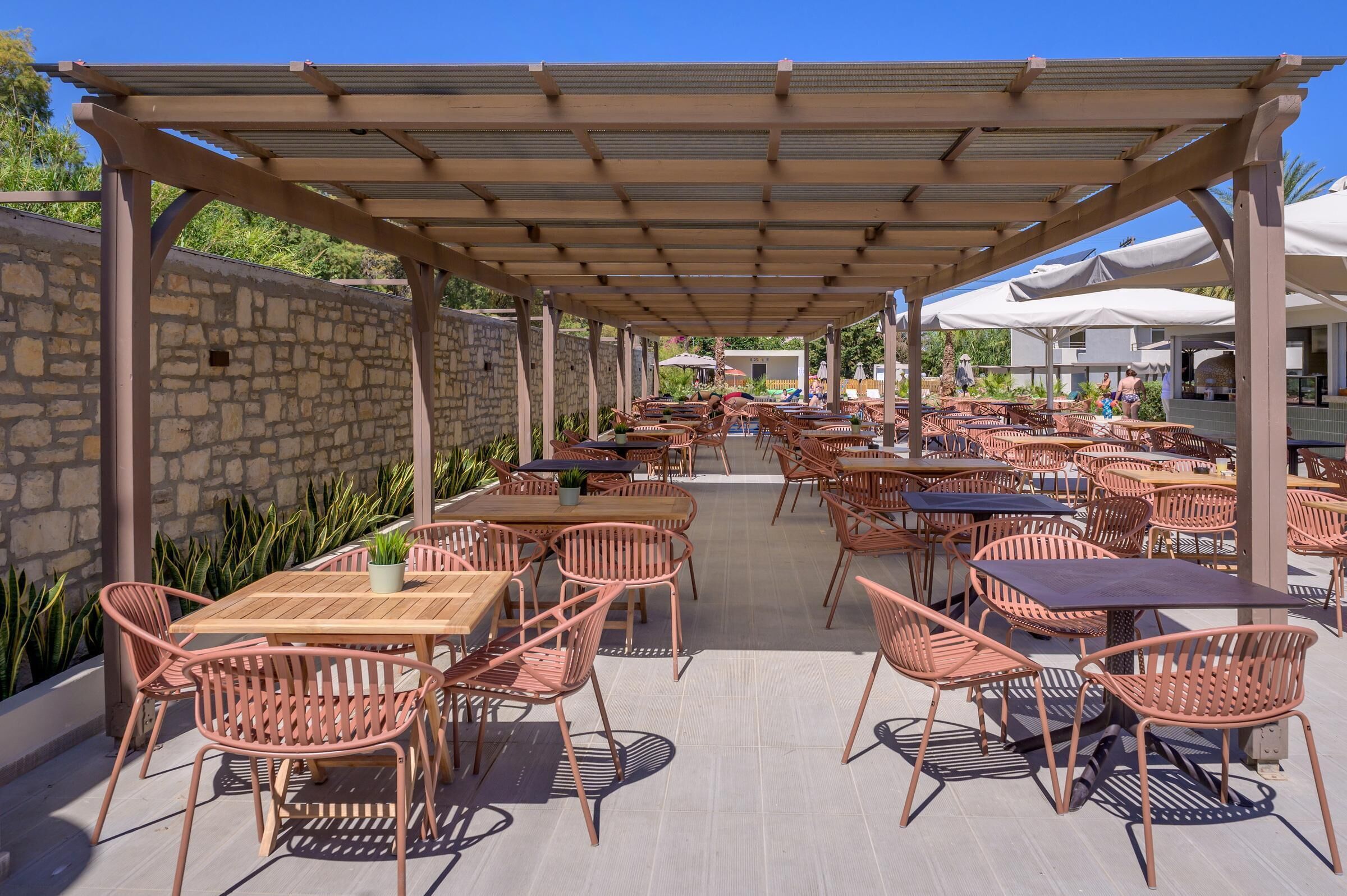 An outdoor patio with wooden tables and chairs, a stone wall, and a pergola.