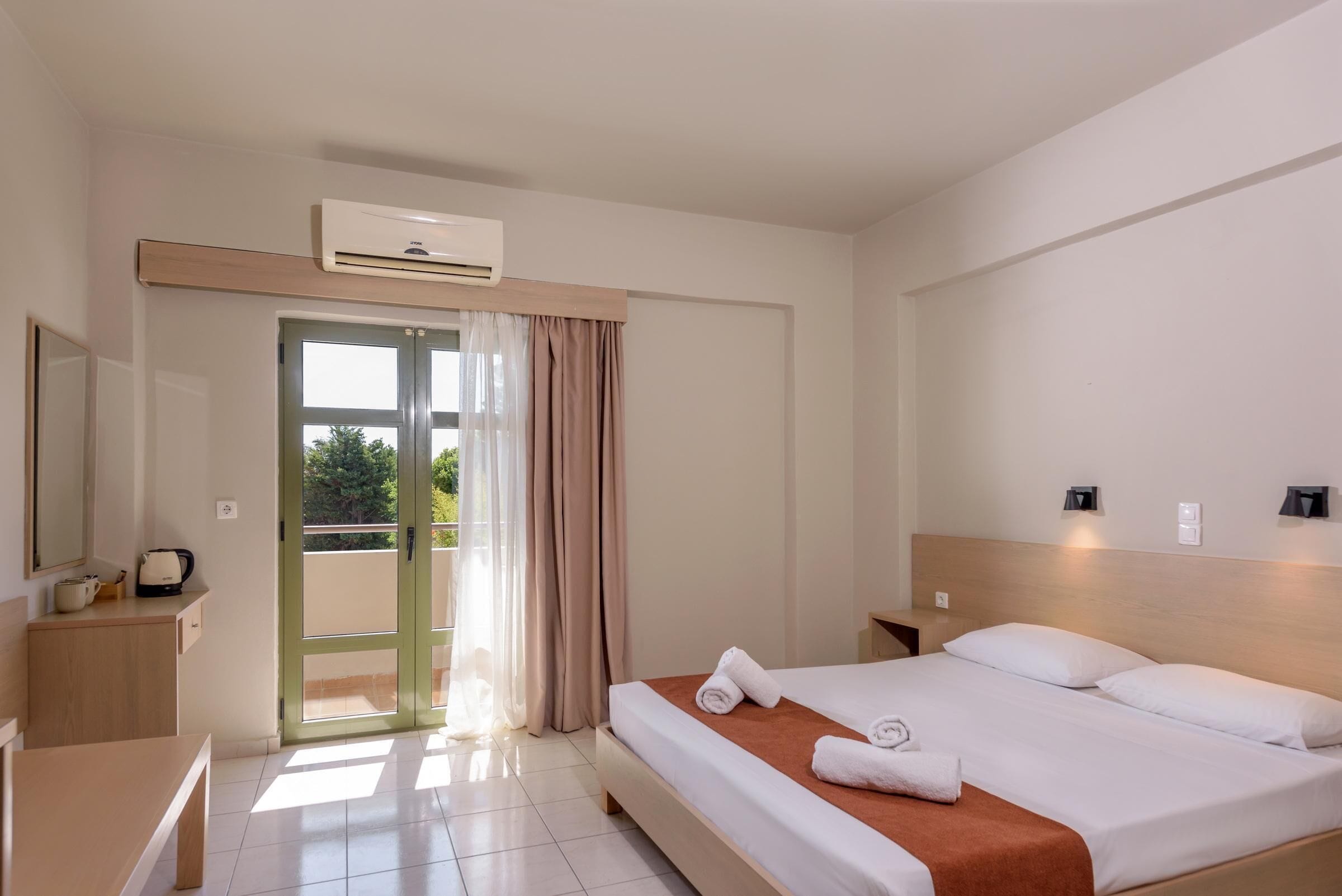 A hotel room with a bed, a desk, a chair, a small table, an air conditioning unit, a nightstand, and a window with curtains.