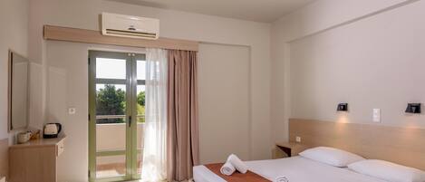 A hotel room with a bed, a desk, a chair, a small table, an air conditioning unit, a nightstand, and a window with curtains.