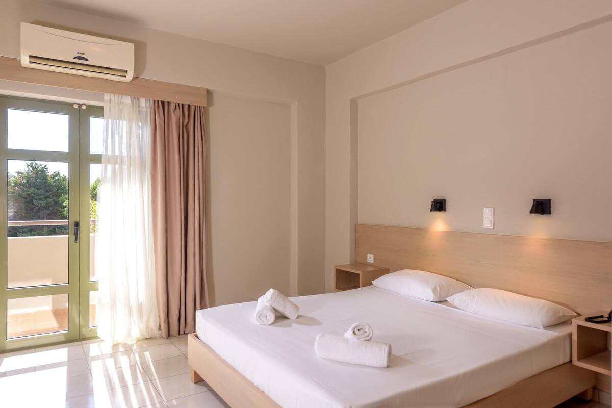 A hotel room with a bed, two bedside tables, a nightstand, an air conditioning unit, and a sliding glass door with curtains.