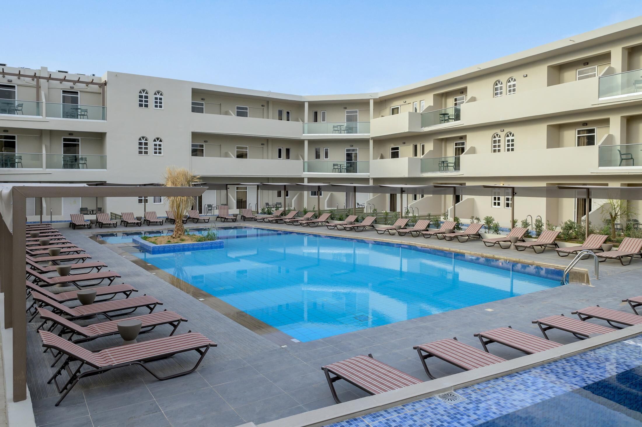 A pool area with sun loungers and a clear blue pool, surrounded by a multi-story building with balconies.