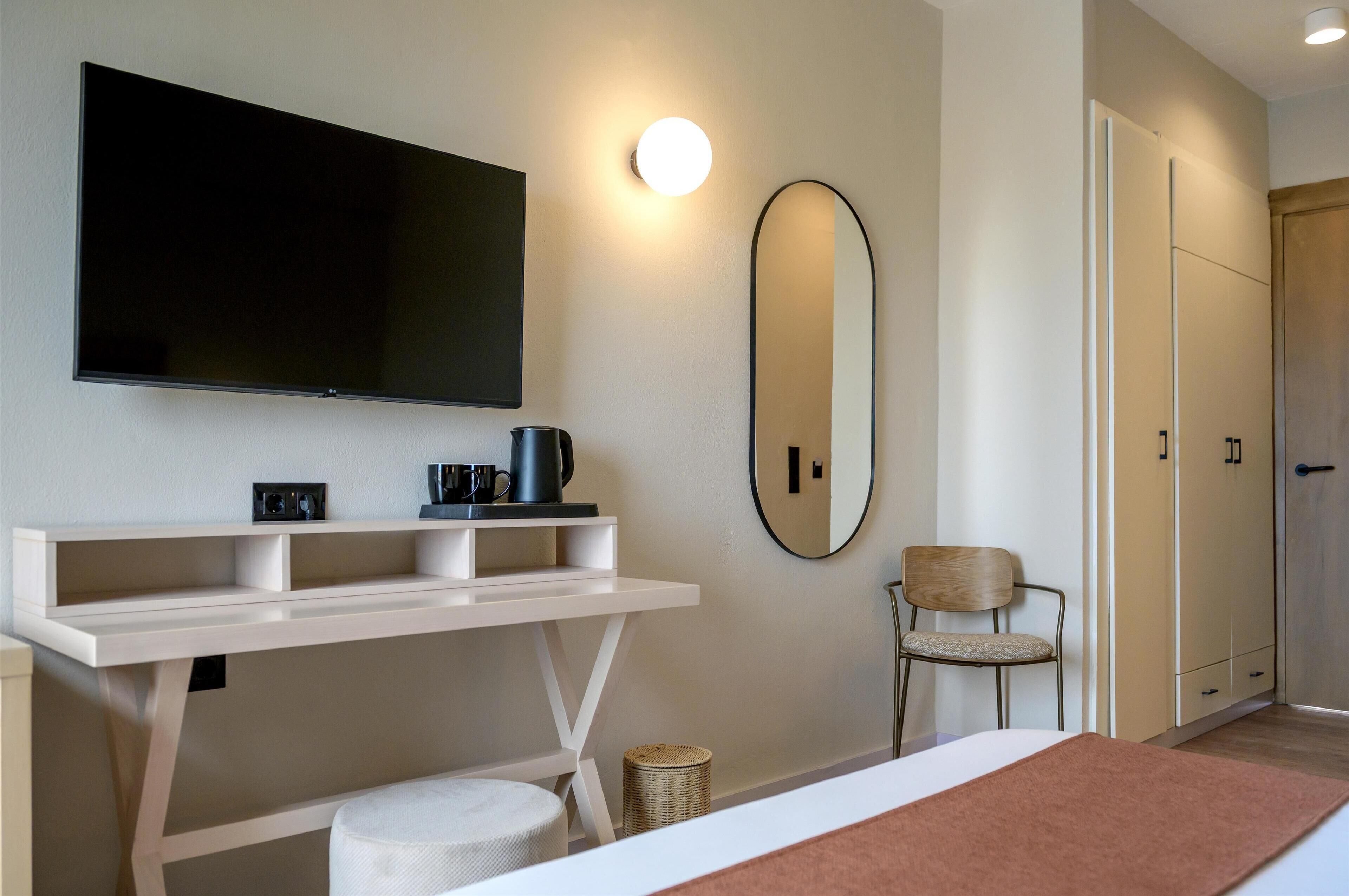 A modern hotel room with a flat-screen TV, a white desk with shelves, a round mirror, a chair, and a wardrobe.