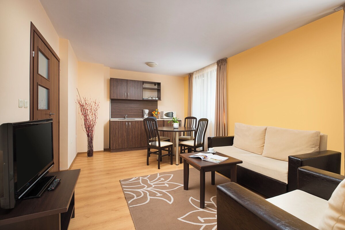 A compact living space with a sofa, a coffee table, a TV, a dining table with chairs, a kitchen area, and a decorative plant.