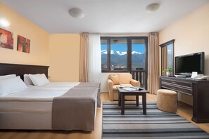 A hotel room with a bed, a desk, a chair, a TV, and a view of mountains. - Royal Park Hotel and Apartments (Bansko)