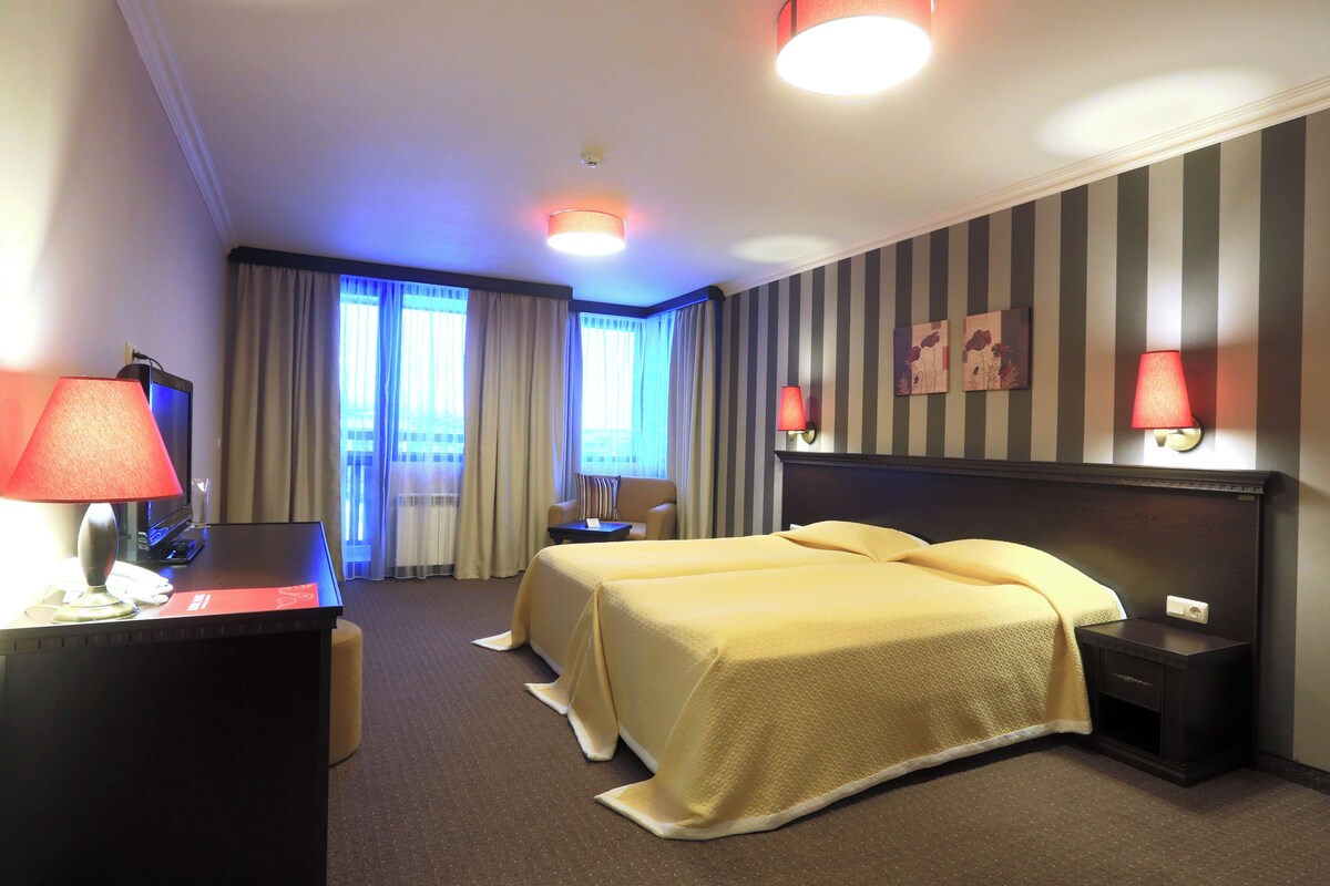 A hotel room with two beds, a desk with a lamp, a chair, a nightstand, and a window with curtains.