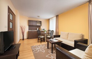 A compact living space with a sofa, a coffee table, a TV, a dining table with chairs, a kitchen area, and a decorative plant. - Royal Park Hotel and Apartments (Bansko)