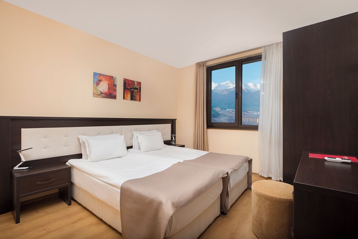 A hotel room with a large bed, a nightstand, a desk, and a view of mountains.