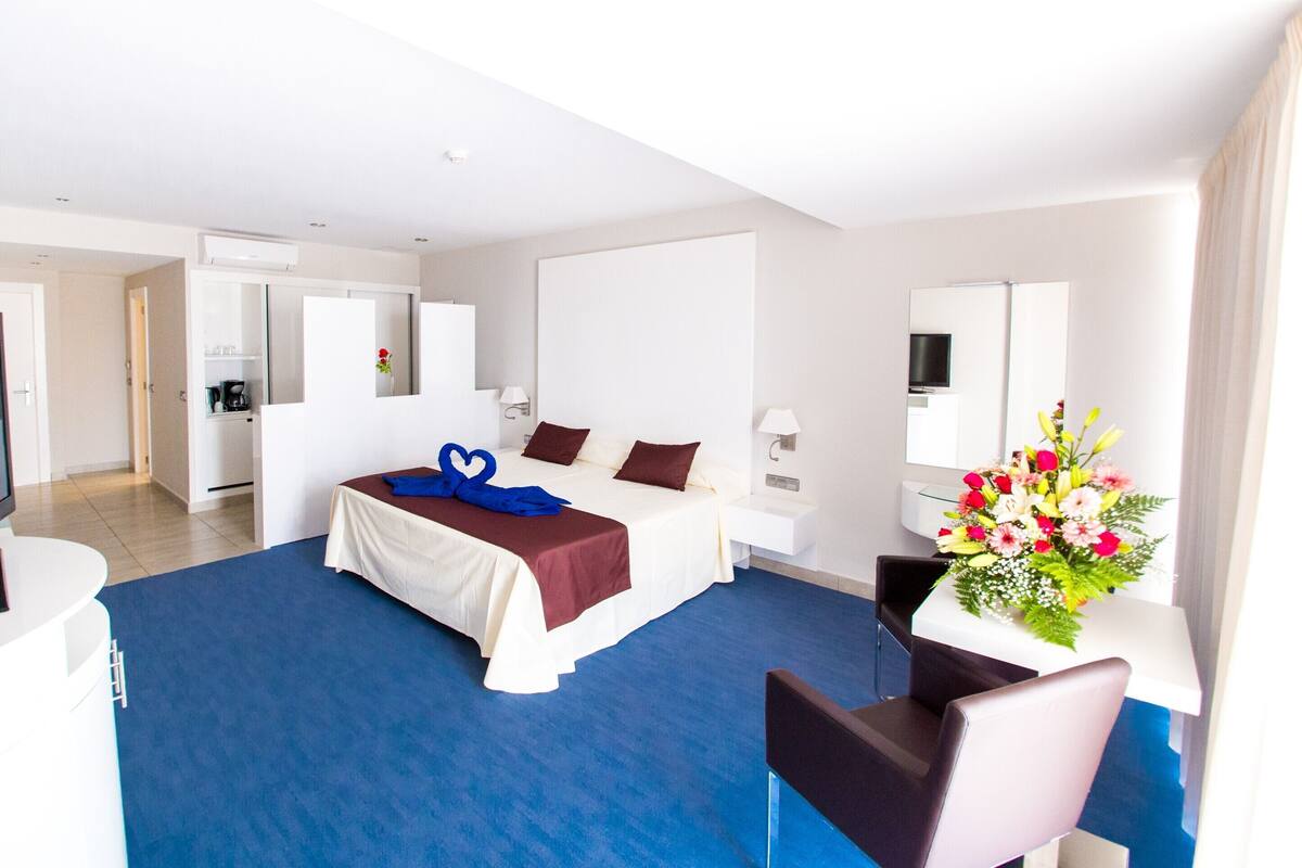 Ein Hotelzimmer mit Bett, Schreibtisch mit Fernseher, Stuhl, Schreibtischlampe und Blumen.
