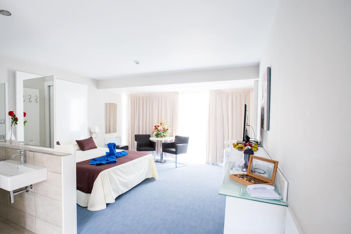 Ein Hotelzimmer mit einem Bett, einem Schreibtisch, einem Stuhl, einem kleinen Tisch mit Blumen in einer Vase, einem Spiegel und einem Fenster mit Vorhängen.