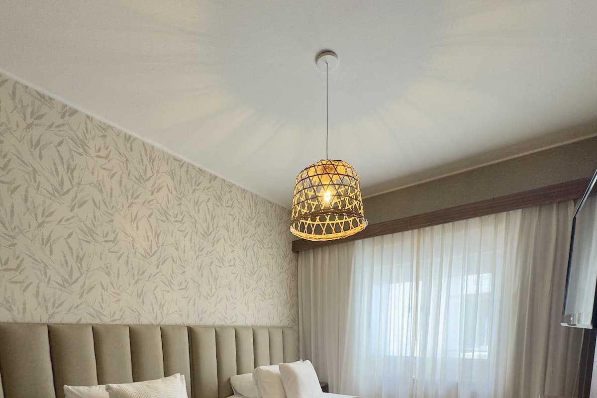 A hotel room with two beds, a large window with sheer curtains, a hanging light fixture, and a wooden floor.