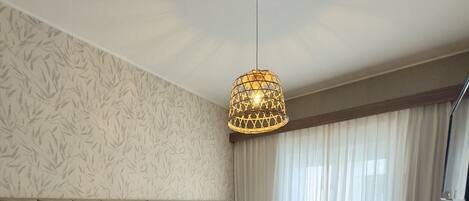 A hotel room with two beds, a large window with sheer curtains, a hanging light fixture, and a wooden floor.