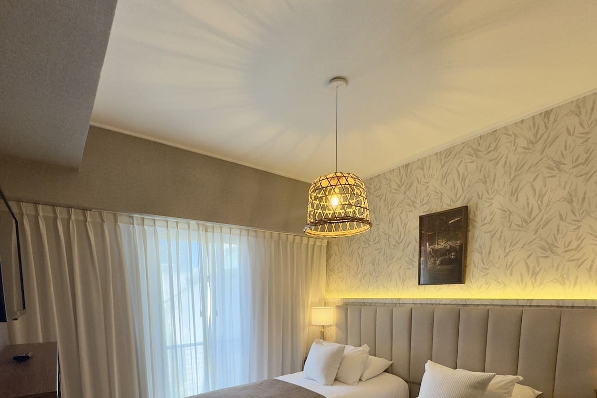 A hotel room with two beds, a wooden floor, a large window with curtains, a hanging light fixture, and a wall-mounted picture.