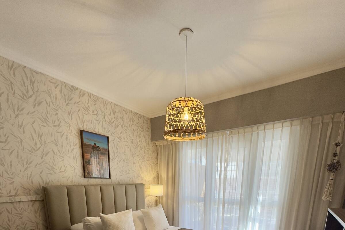 A bedroom with a large bed, a headboard, a nightstand, a lamp, a TV stand, and a window with curtains.