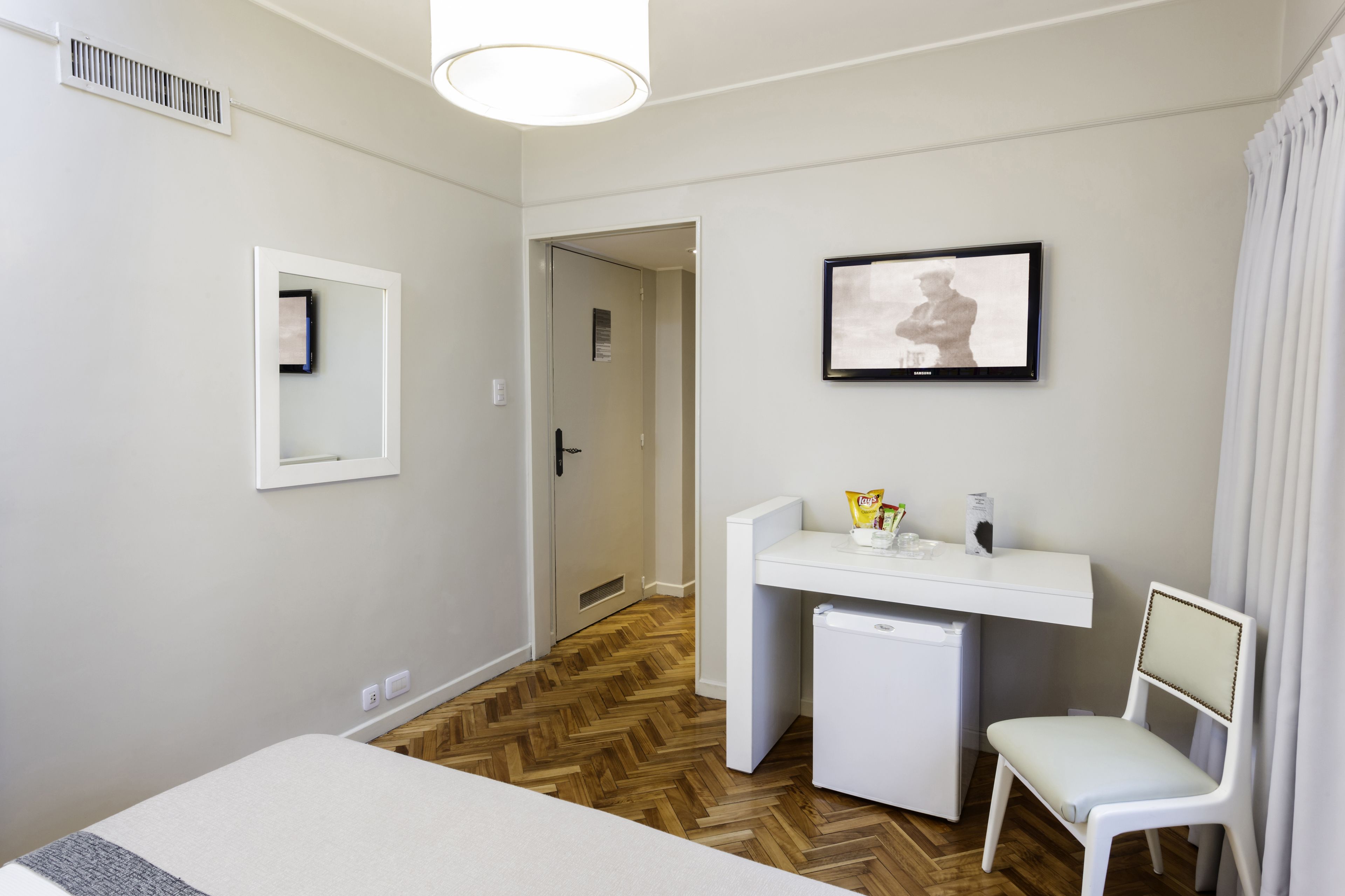 A hotel room with a bed, a desk with a chair, a small refrigerator, a television, and a door leading to another room.