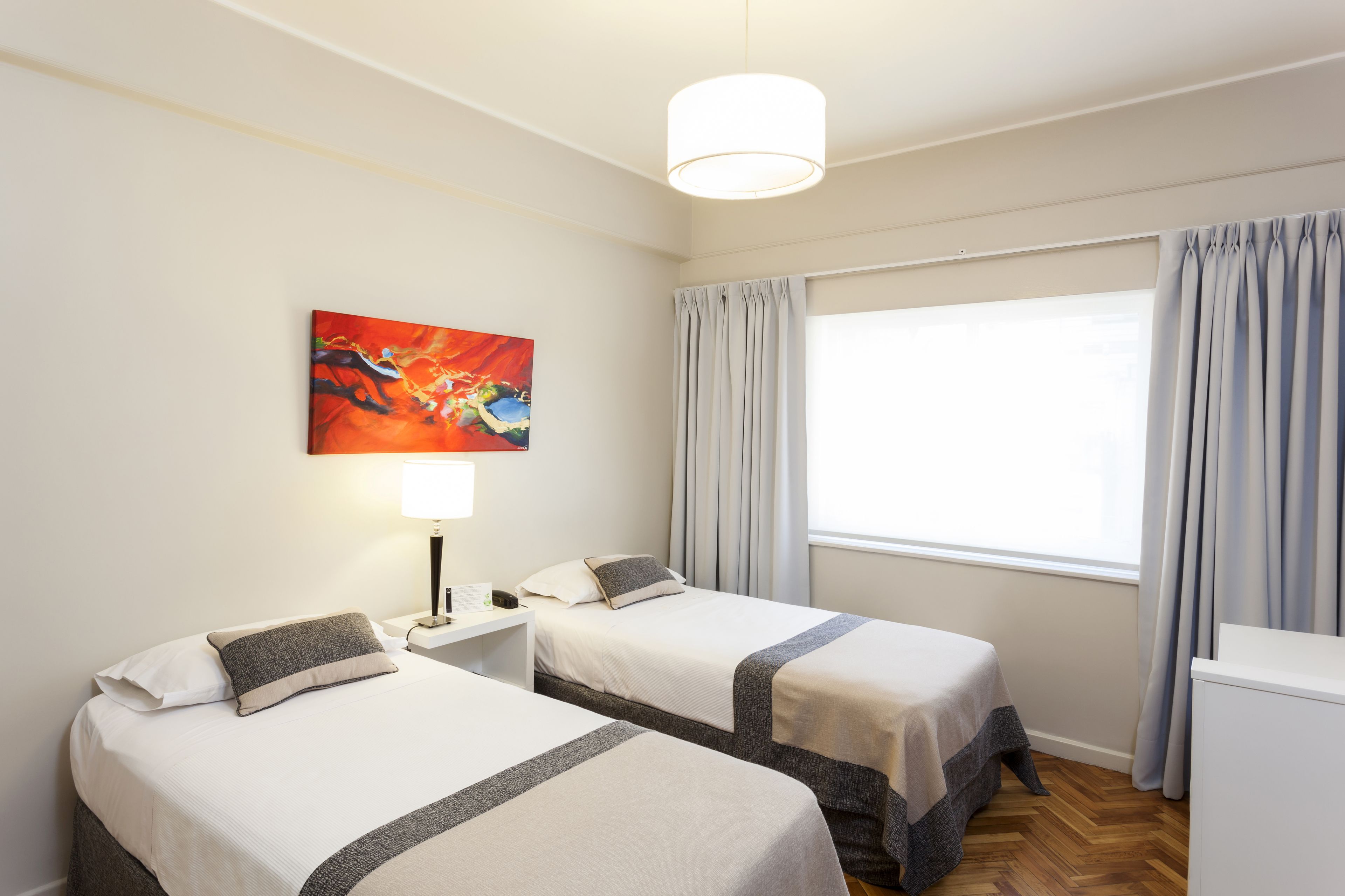 A hotel room with two beds, a painting on the wall, a desk with a lamp, and a window with curtains.
