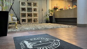 Un vestíbulo de hotel con mostrador de recepción, un aparador decorativo con puertas de cristal, felpudo negro con logotipo e iluminación empotrada en el techo.