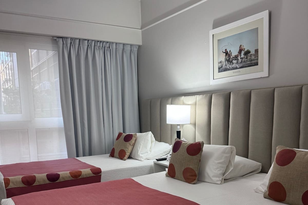 A hotel room with two beds, a headboard, a nightstand, a lamp, and a window with curtains.