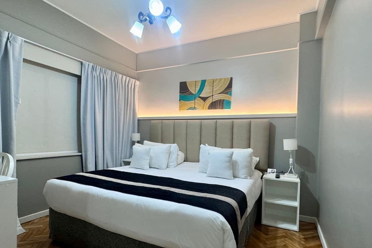 A modern hotel room with a large bed, a bedside table, a nightstand, a ceiling light fixture, and a window with curtains.