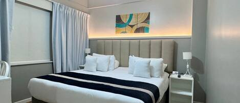 A modern hotel room with a large bed, a bedside table, a nightstand, a ceiling light fixture, and a window with curtains.